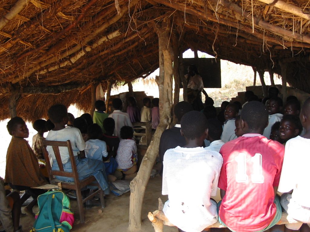 Children sit with backs to us in a classroom with simple pitched thatched roof with no walls