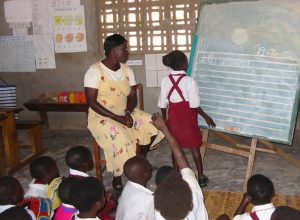 Teacher (seated) and child (standing) in front of blackboard. Other children sitting on floor facing board