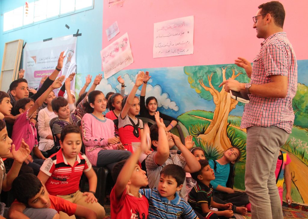 Group of girls and boys in Gaza, half have hands raised, smiling, looking at adult male (Ayman) who is standing on right of image talking/asking question. Colourful landscape mural painted on teh wall.