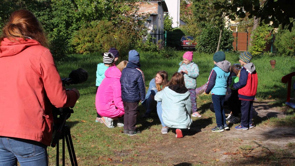 Woman with camera standing on left, viewing a group of small children in a circle.