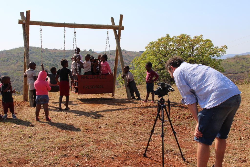 Wheelchair accessible swing, children on it. Cameraman in foreground.