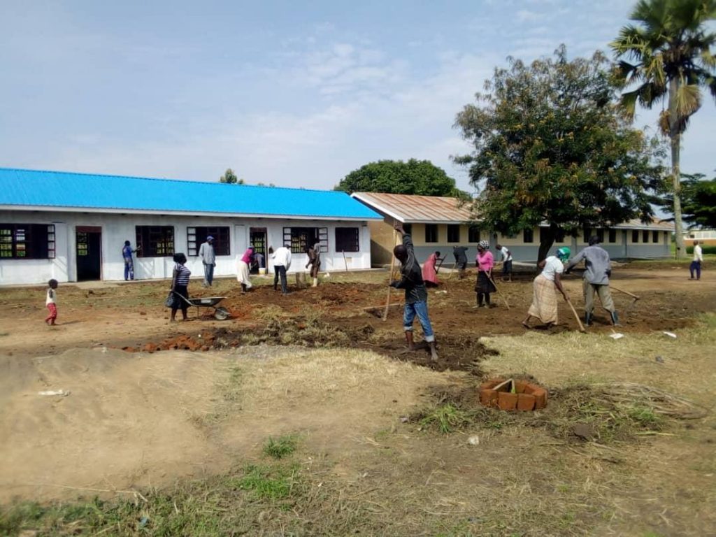 A group of men and women are using hand-tools to dig and level the ground in front of two school buildings.