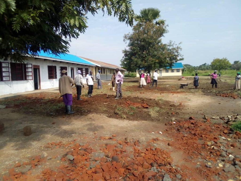 A group of men and women are using hand-tools to dig and level the ground in front of two school buildings.