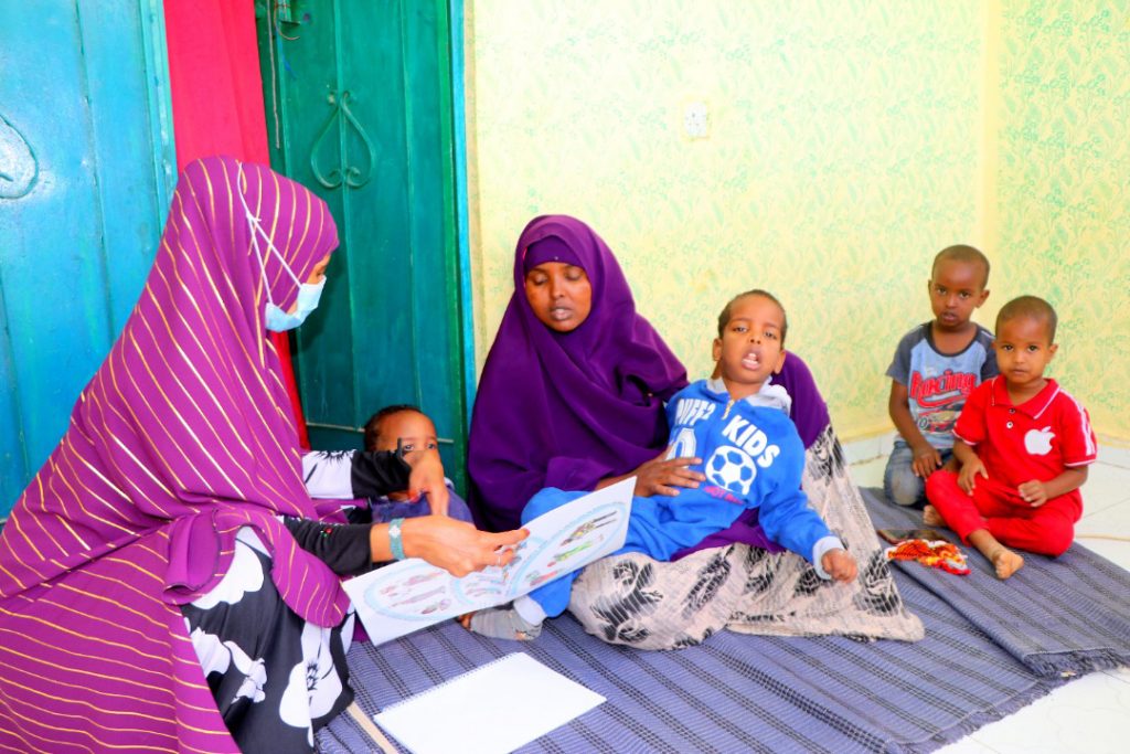 A mother and her children are sat on a mat on the floor and are being shown some of the home learning materials by a volunteer distributor