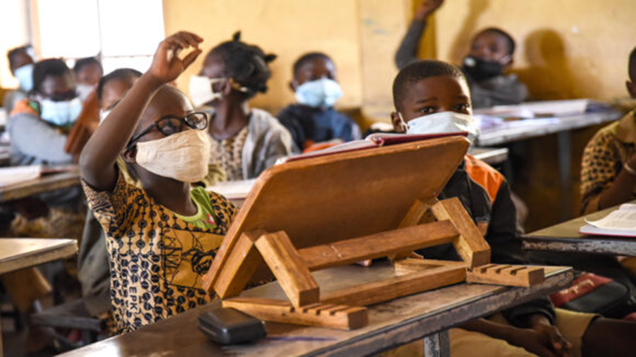 A girl wearing glasses and a face mask sits in a class with other children. She raises her right hand. In front of her on the desk is a raised sloping book stand.