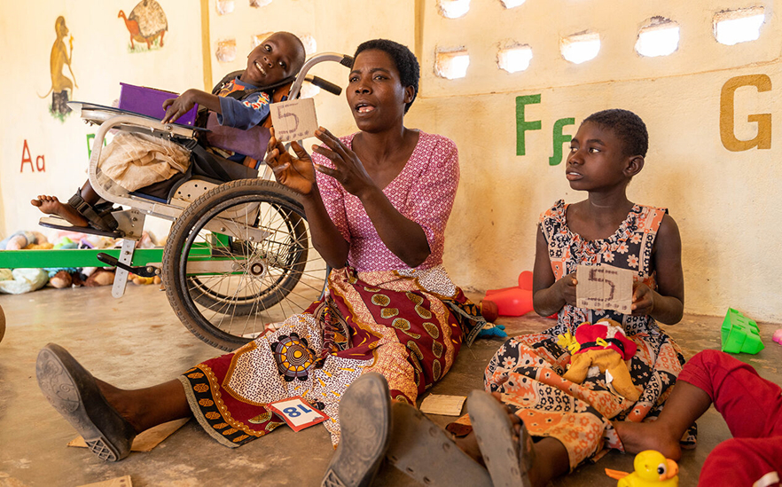 In the middle, a woman sits on the floor holding a card saying '5'. On the left a girl sits in a wheelchair, On the right a girl sits on the floor also holding a card that says '5'. The wall behind has the alphabet painted on.