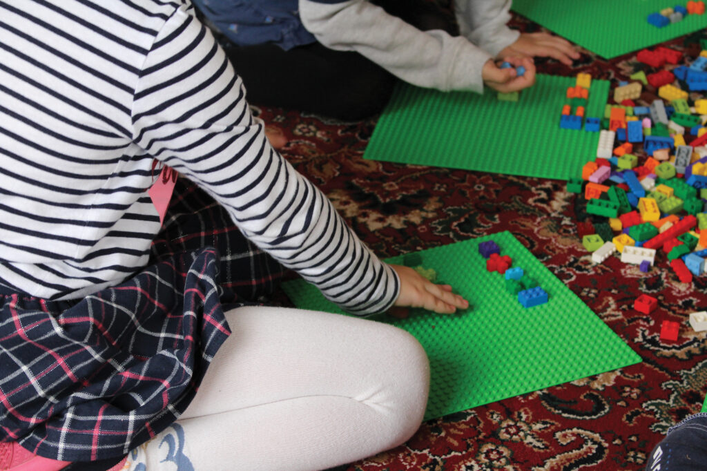 Two young children are sitting on the floor. We only see part of their bodies, arms and legs. They have a pile of Lego building blocks in front of them. Each child is putting blocks onto a Lego base plate.
