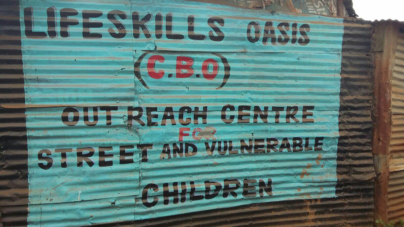 Image shows a corrugated iron fence painted with a blue square. The text drawn over the square says: Life Skills Oasis (C.B.O.) outreach centre for street and vulnerable children.