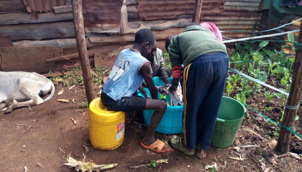 Three boys/young men are gathered around a large blue bucket. One boy is sitting on a barrel, one is crouching, one is standing and bending down. They appear to be washing clothes in the bucket.