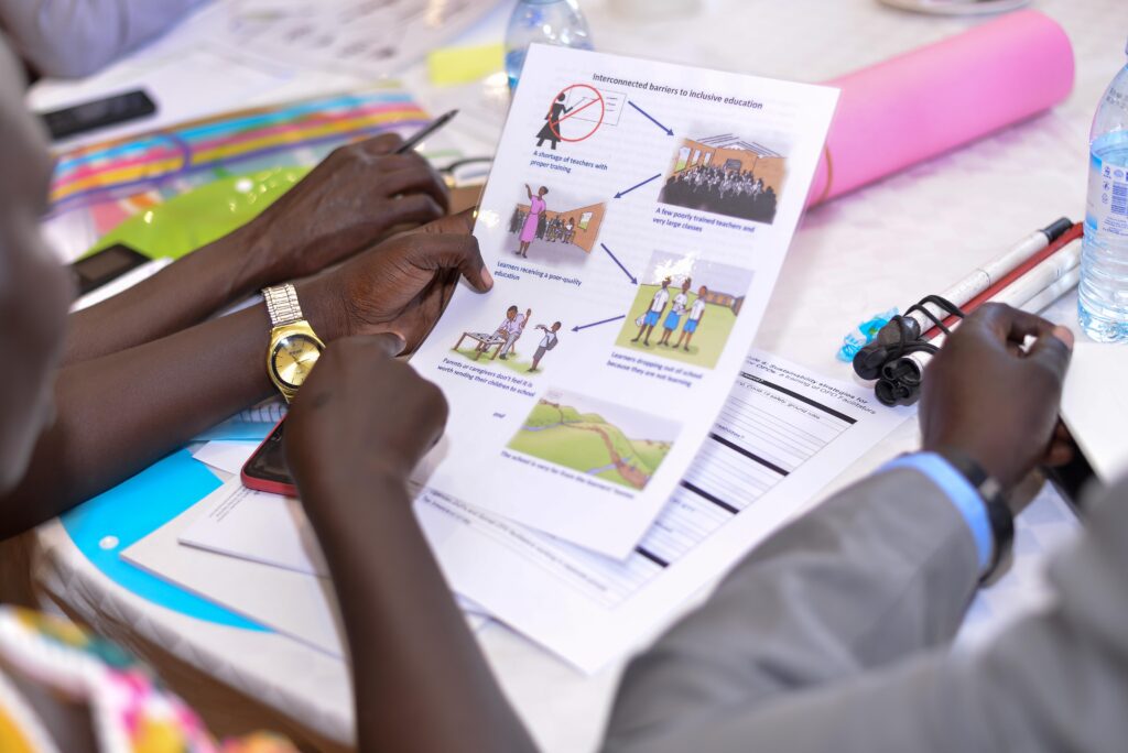 A person seated at a table (we only see arms and hands) is holding a sheet of paper in their left hand. The paper has 6 small illustrations on it. There is another person (we see arms and hands only) sitting to the left and to the right. They appear to be discussing the illustrations.