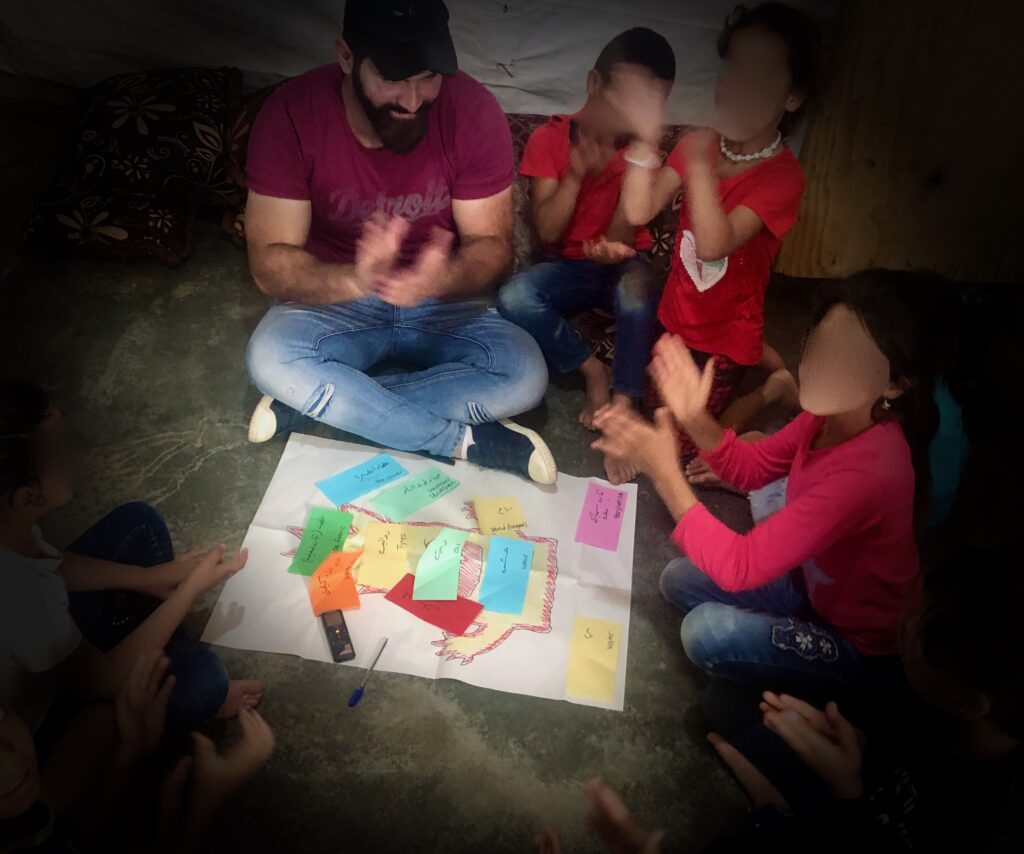 A man sits on the ground in a circle with 6 children. They are all clapping. On the floor in the middle of the circle is a post with a monster drawn on it. Stuck on top of the monster are different coloured post it notes with words on.