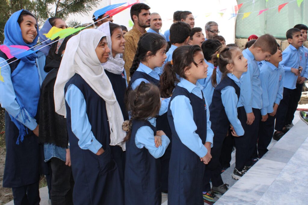 Approximately 15 girls and boys in blue school uniform are standing in rows facing and looking intently at what may be someone or something on a stage (not visible). An adult stands behind them.