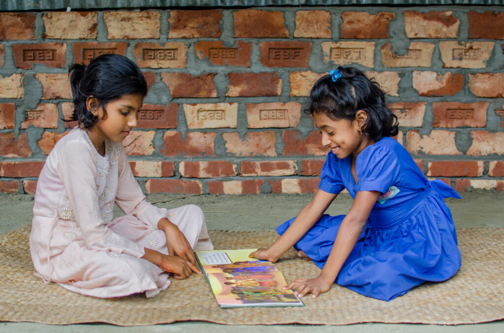Two girls sit on the floor facing each other. Between them is an open illustrated story book. One girl is smiling.