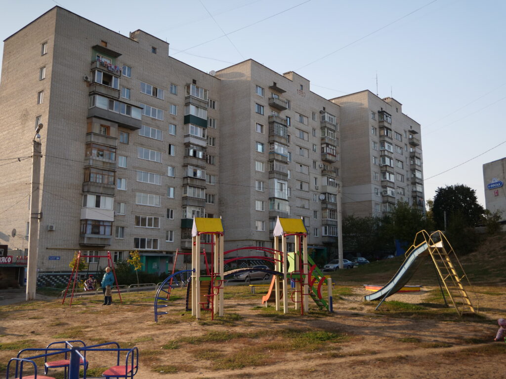An outdoor playground is located o na grass area in front of 3 large apartment blocks. The playground has colourful apparatus like slides, swings, climbing frames and a roundabout. A small child is sitting on a swing, with an adult standing close by.