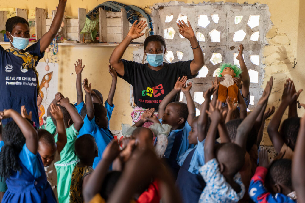 A female teacher is standing in a classroom. Her arms are raised above her head, like she is exercising or waving. In front of her are approximately 15 small children who also have their arms raised. Another woman stands to the side, again with arms raised. Both adults wear COVID-19 face masks.