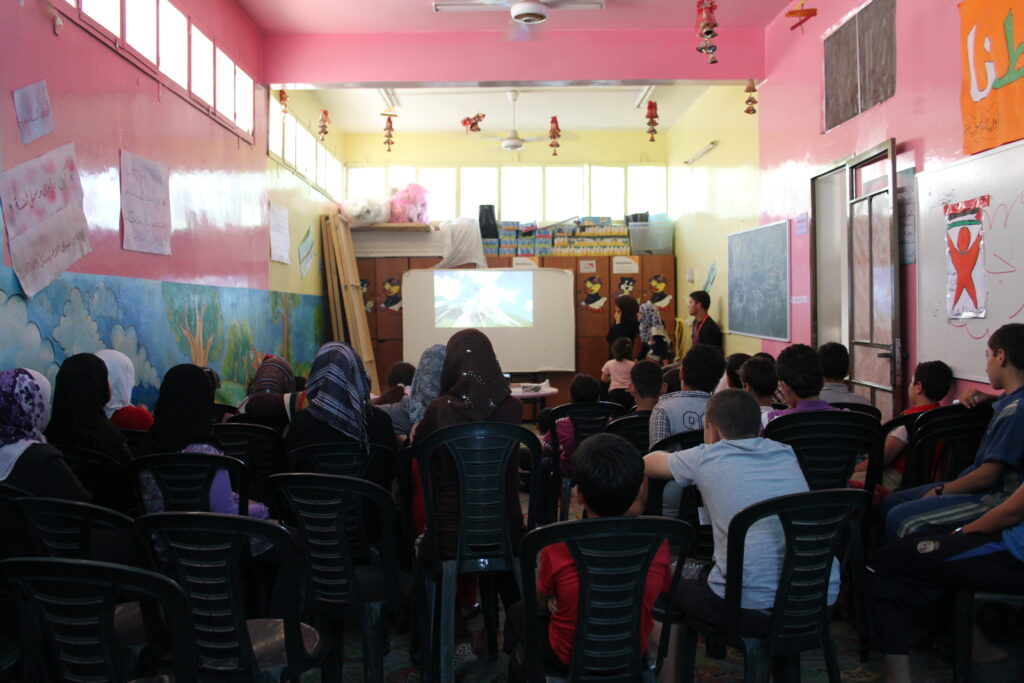 Approximately 30 girls and boys are sitting on chairs in rows. They have their backs to the camera and are watching a film on a screen at the front of the room. The room is brightly decorated and may be a classroom.