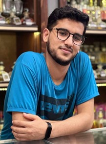 Osama Abu Saifa smiles at the camera as he leans on a counter. He has his arms crossed and is wearing a blue t shirt. There are trophies on the shelves behind him. 