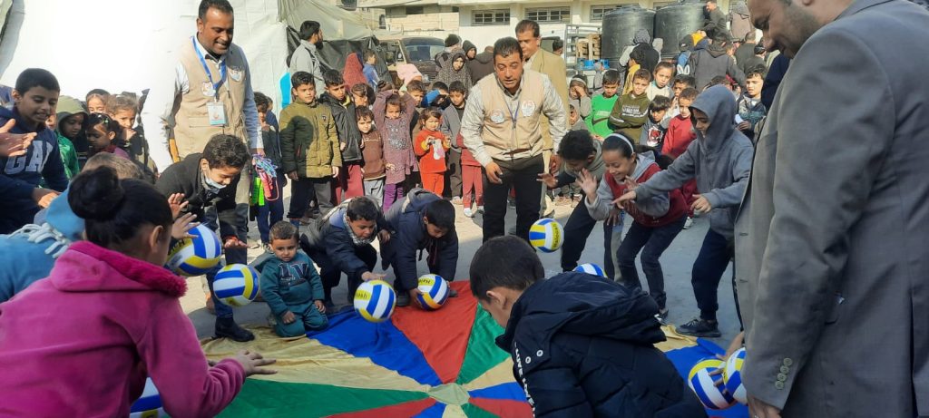 Many children and a few adults playing with a rainbow coloured sheet of cloth and some balls