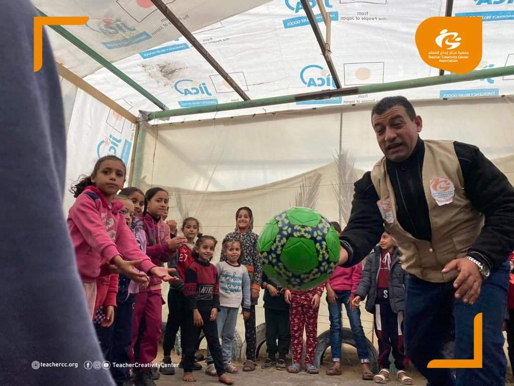 A man hoding a ball, with children around him. They are inside a refugee tent.