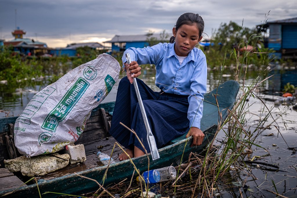 A girl sits in a boat on a river with a large white plastic bag next to her. She is using a litter picker to grab a plastic bottle from the water.