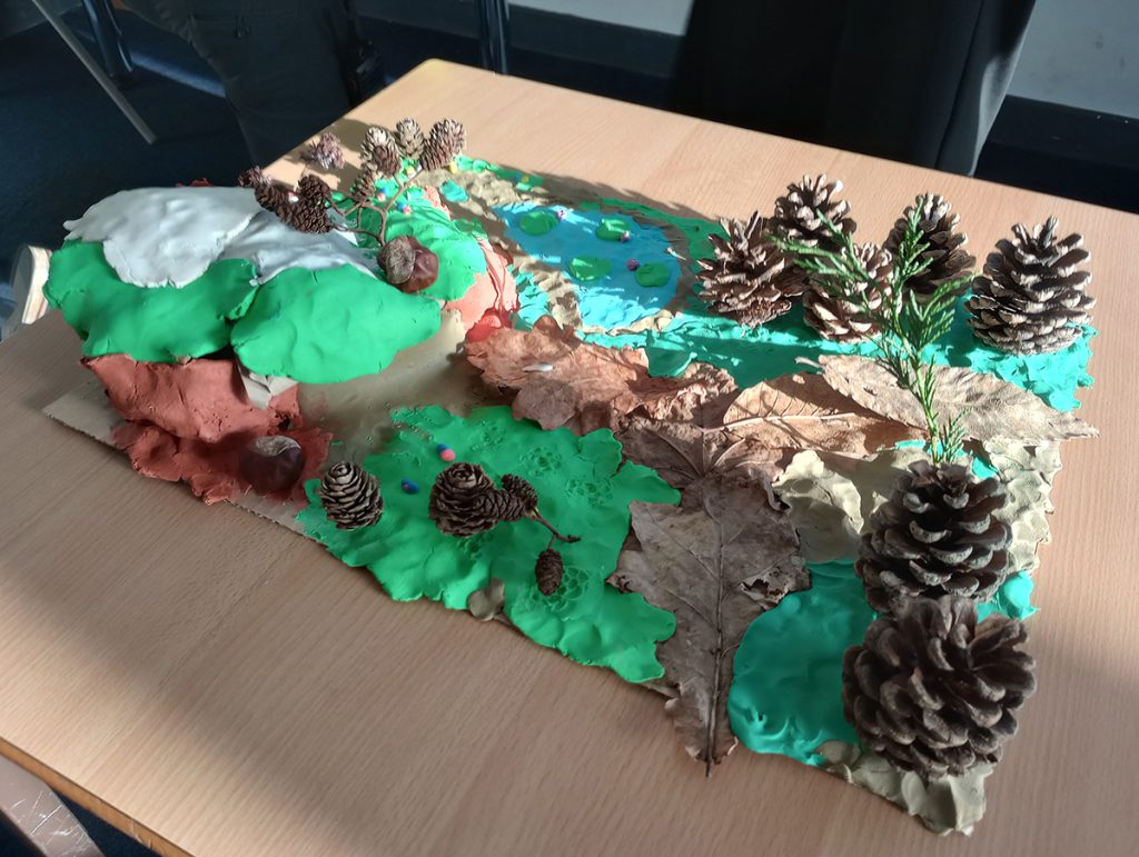 A landscpe made of clay in green, grey, blue and brown shows a hill, a pond and pathway through pinecones used to represent trees in a woodland.  