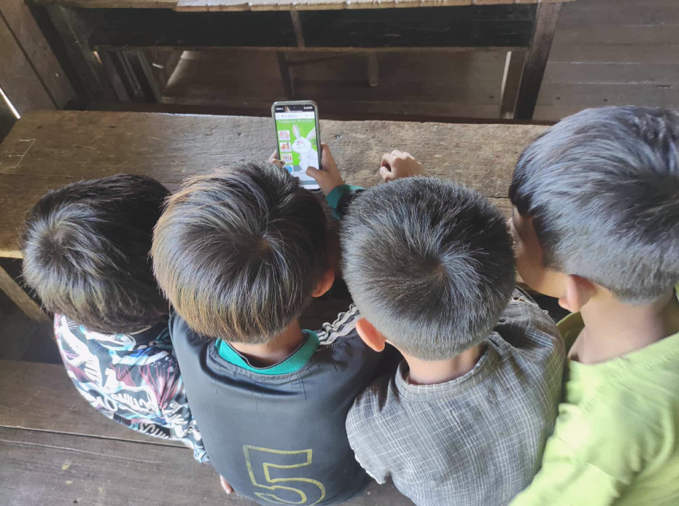 The view is of the back of the heads of four boys sat together at a school desk as they all look at the screen of a mobile phone.