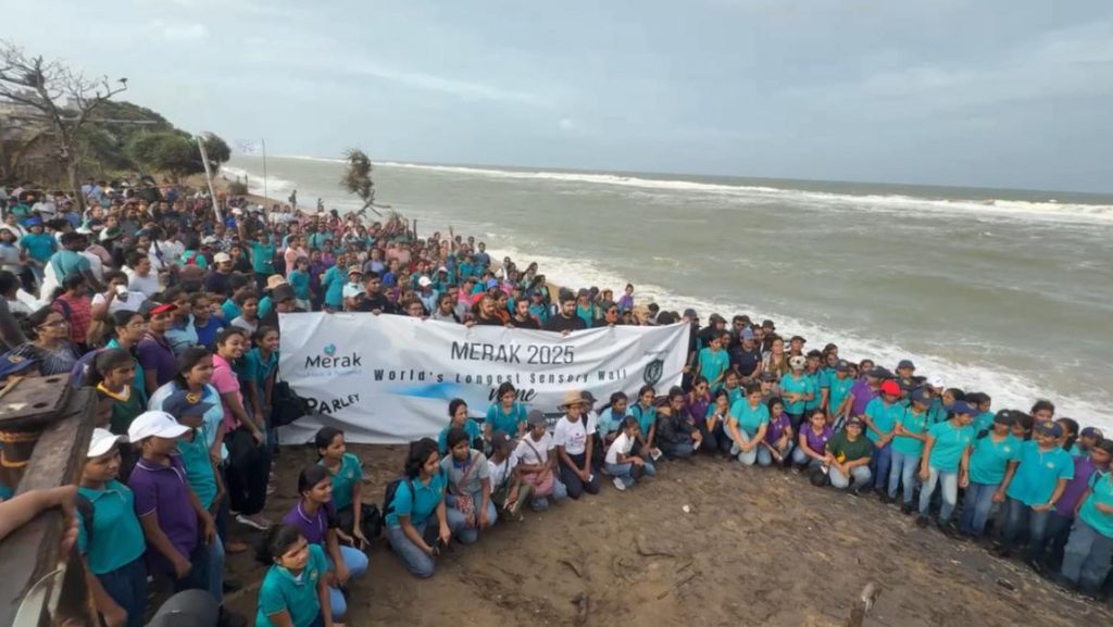 A large crowd of people stand and sit on a beach as the waves roll in. They are all posing for the photograph and holding a banner advertising Merak and the world's longest sensory wall.